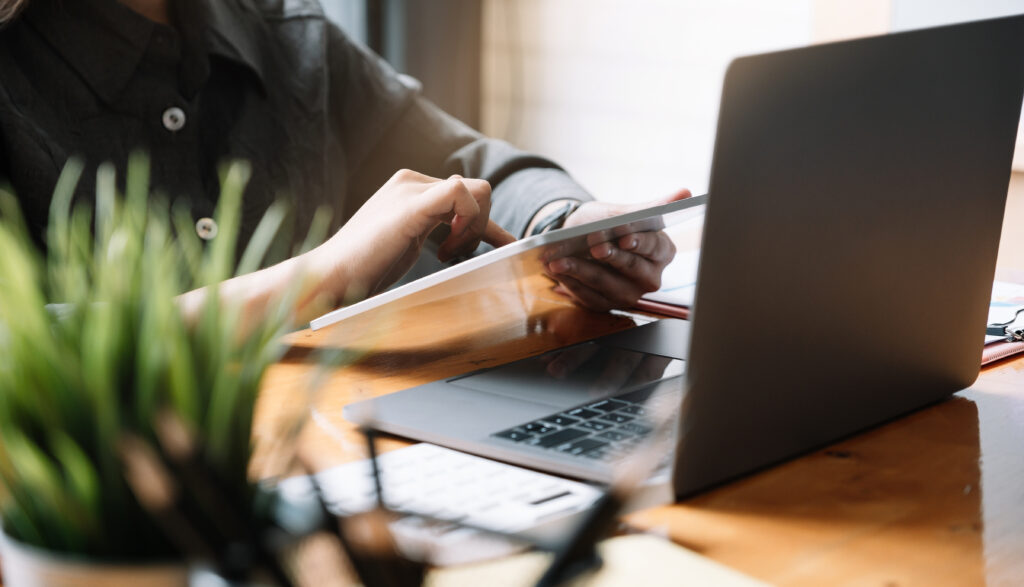 Person am Schreibtisch mit Tablet in der Hand vor einem Laptop
