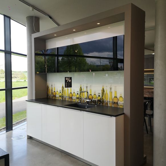 Ausstellungsküche mit Küchenzeile bestehend aus weißen hochglanz Unterschränken, Oliven-Glas-Rückwand und schwarzen Glas-Oberschränken